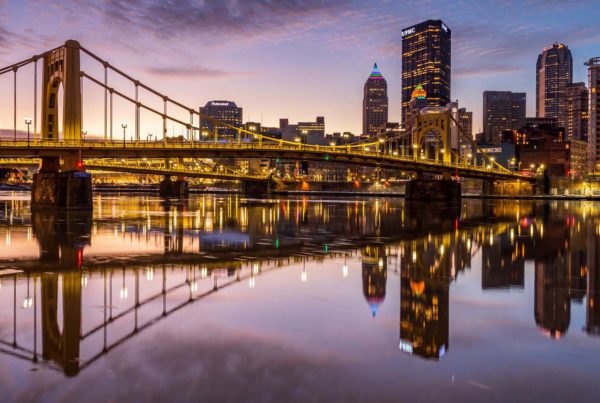 A twilight image of the Allegheny River from the North Side with one of the yellow three sisters bridges spanning the image with the downtown skyline on the far right.