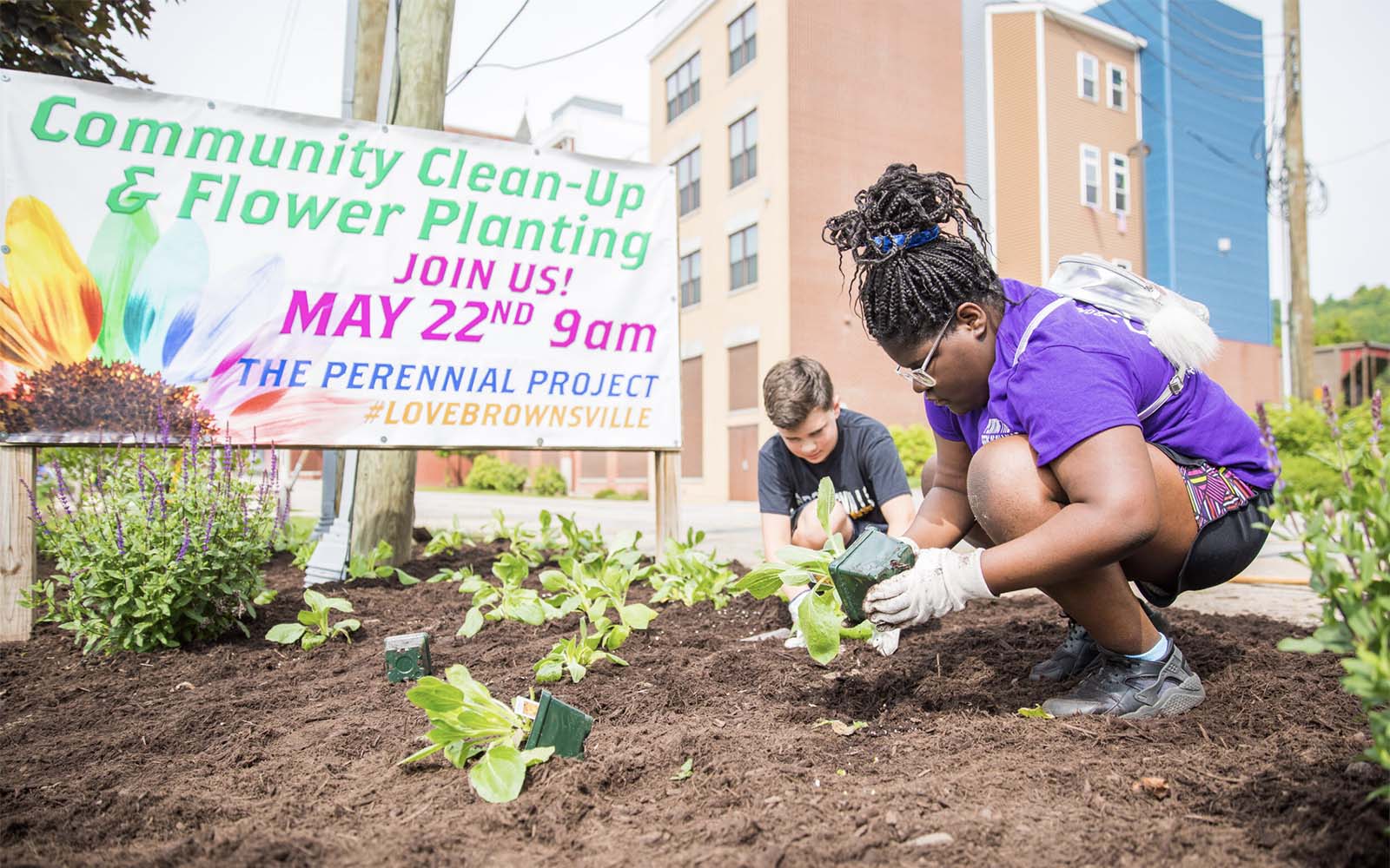 Two people, one dark skinned and one white, crouch down to plant flowers in front of a sign that reads "Community Clean-up and Flower Planting May 22"