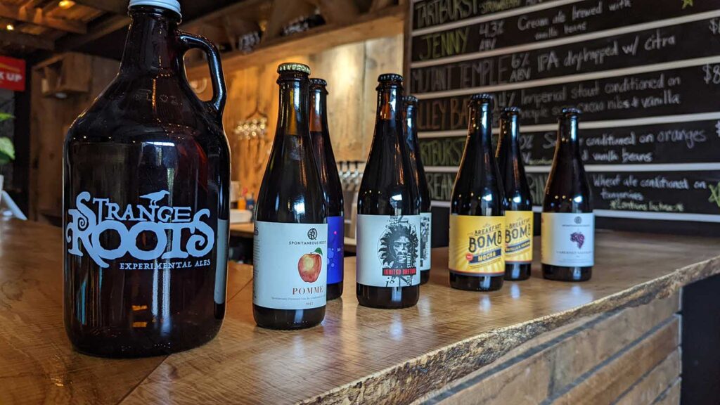 A grower and bottles lined up on a wooden counter with a chalk board of beer listings in the background.