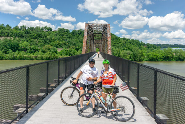 Two women bikers on a converted rail road bridge with blue skies, puffy white clouds, green hills and a healthy green river.