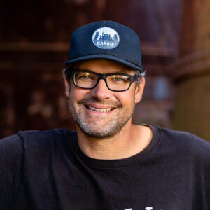 A white man with graying stubble on his chin in a ball cap and glasses smiles for a headshot.