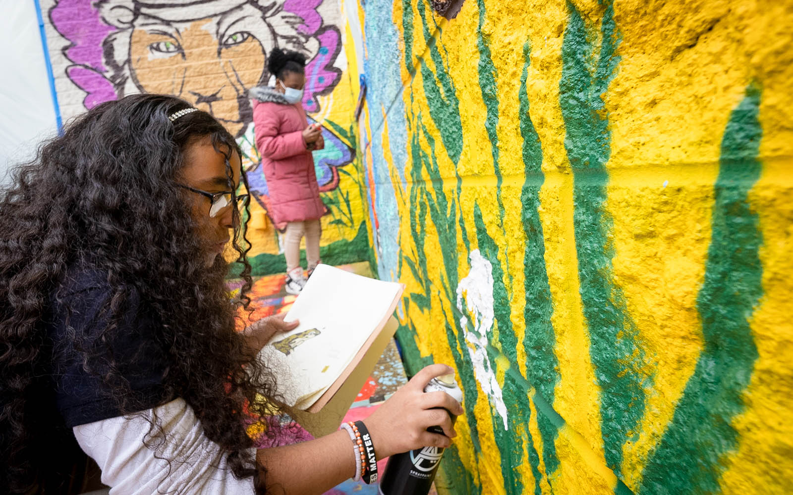 A medium skinned black girl with long curly hair raises a spray can to paint a outdoor mural while another girl paints in the background.