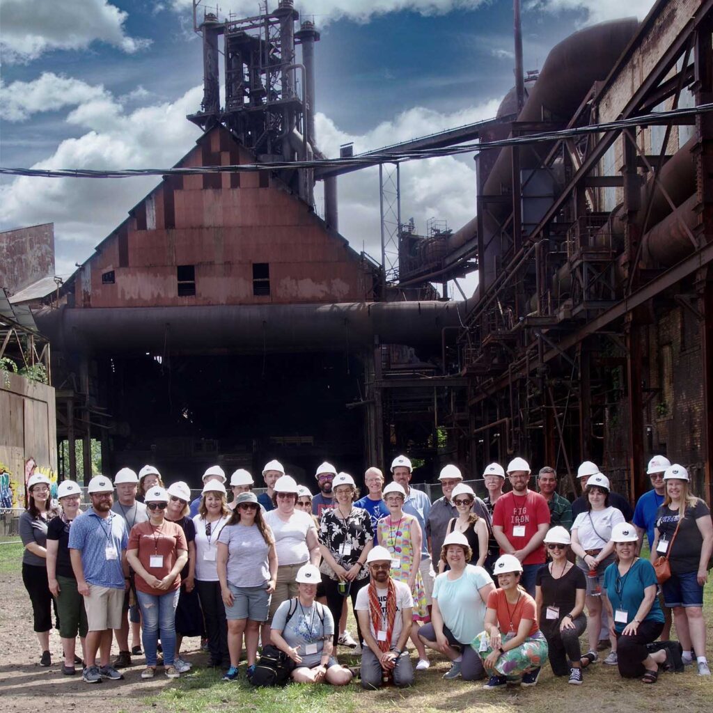 A group of 30 people pose for a photo in front of an industrial structure.