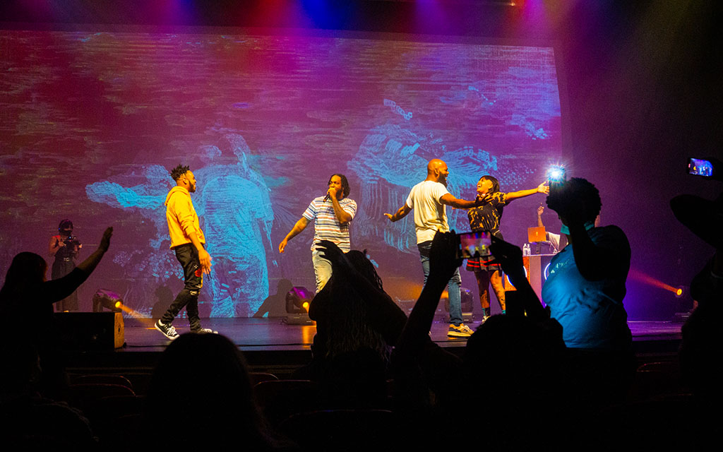 Four black performers dance and sing onstage while a crowd shown in silhouette, appears in the foreground, raising hands and taking photos.