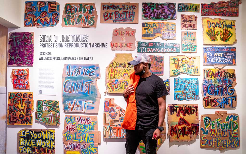 A black man stands in front of a gallery wall, his gaze directed off camera. The artworks are hung salon-style and depict protest signs recreated on cardboard.