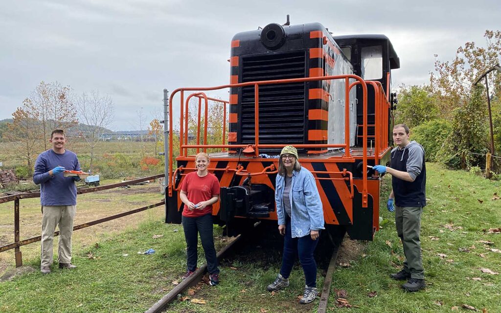 Four volunteers, several with paint or brushes in hand, stand in front of the engine.