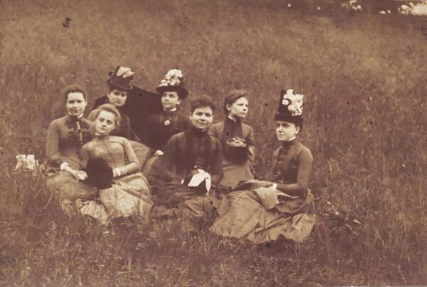 A sepia-toned image of seven young women in high collared dresses lounging in a field of tall grass.