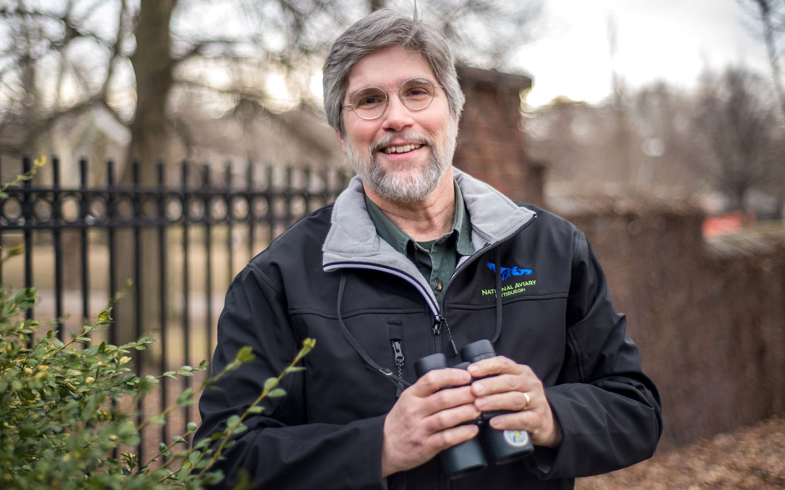 A middle aged white guy with a salt and pepper neatly trimmed beard holds binoculars and smiles gently for the camera.