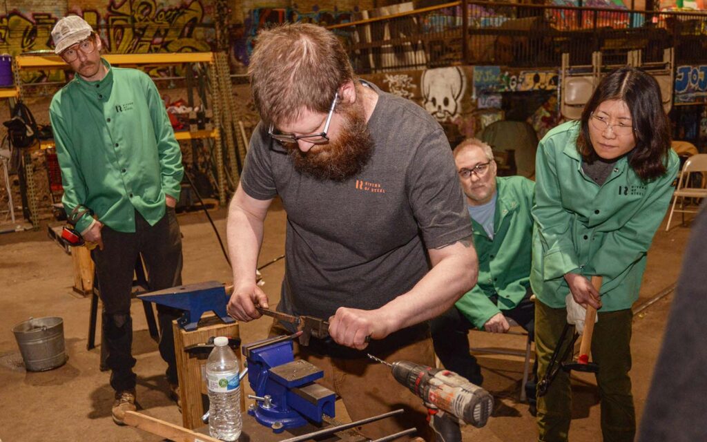 Students in green protective wear gather around the instructor as he bends hot metal at a vice.