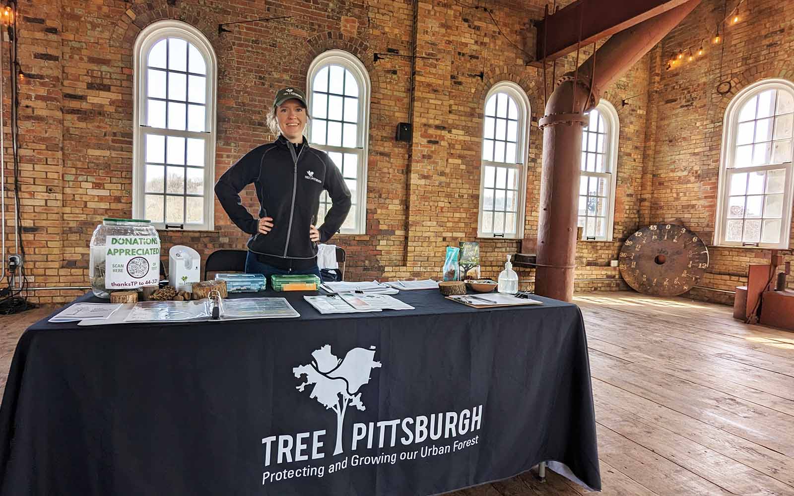 A youthful white woman stands behind a table with a Tree Pittsburgh table covering on it with her hands on her hips, ready to greet visitors with a smile.