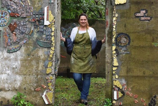 A white woman with reddish hair in jeans and a long sleeve tshirt wearing a smock stands in a concrete doorway that is adorned with mosaic artworks.