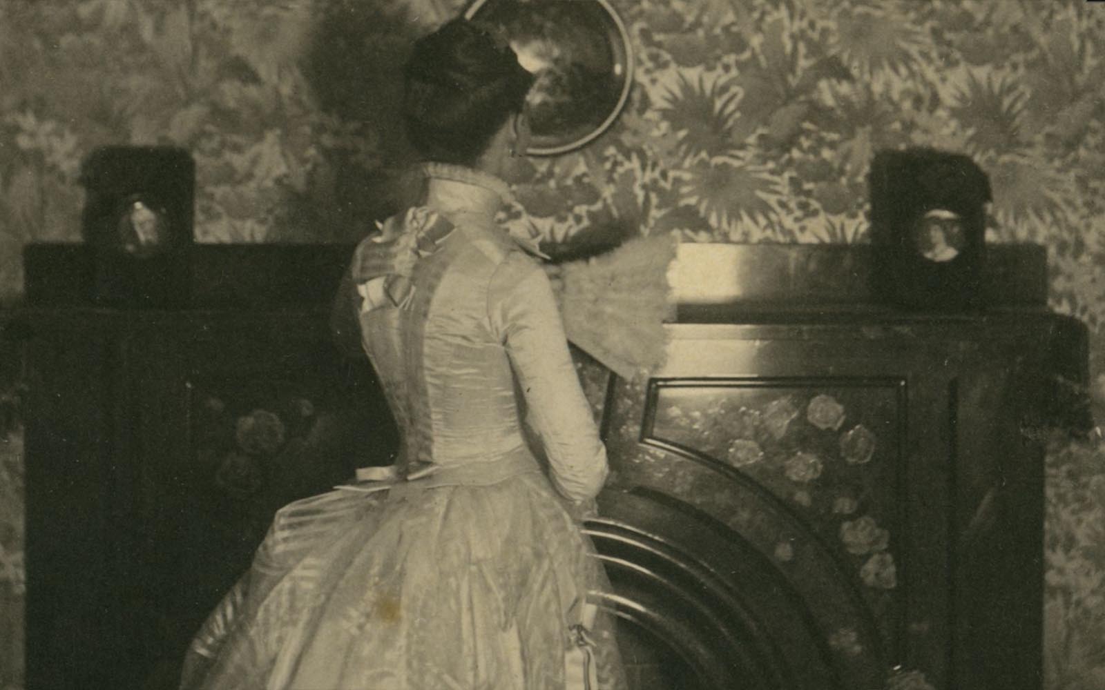 A woman in a white wedding dress with a bustle, high collar, and long sleeve, faces away from the camera.
