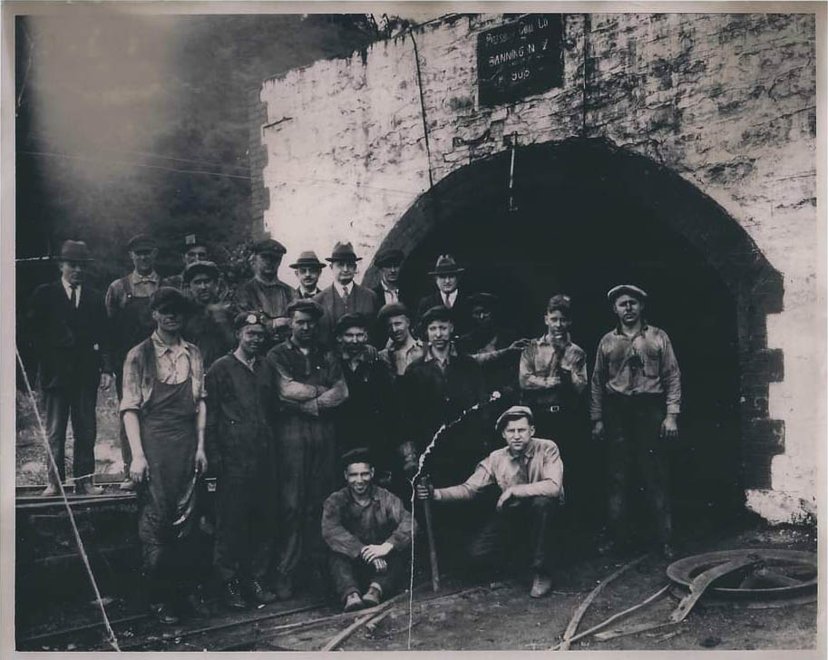 A group of about 20 miners, dirty from their labor, pose for a picture in front of a large arched opening to the mine.