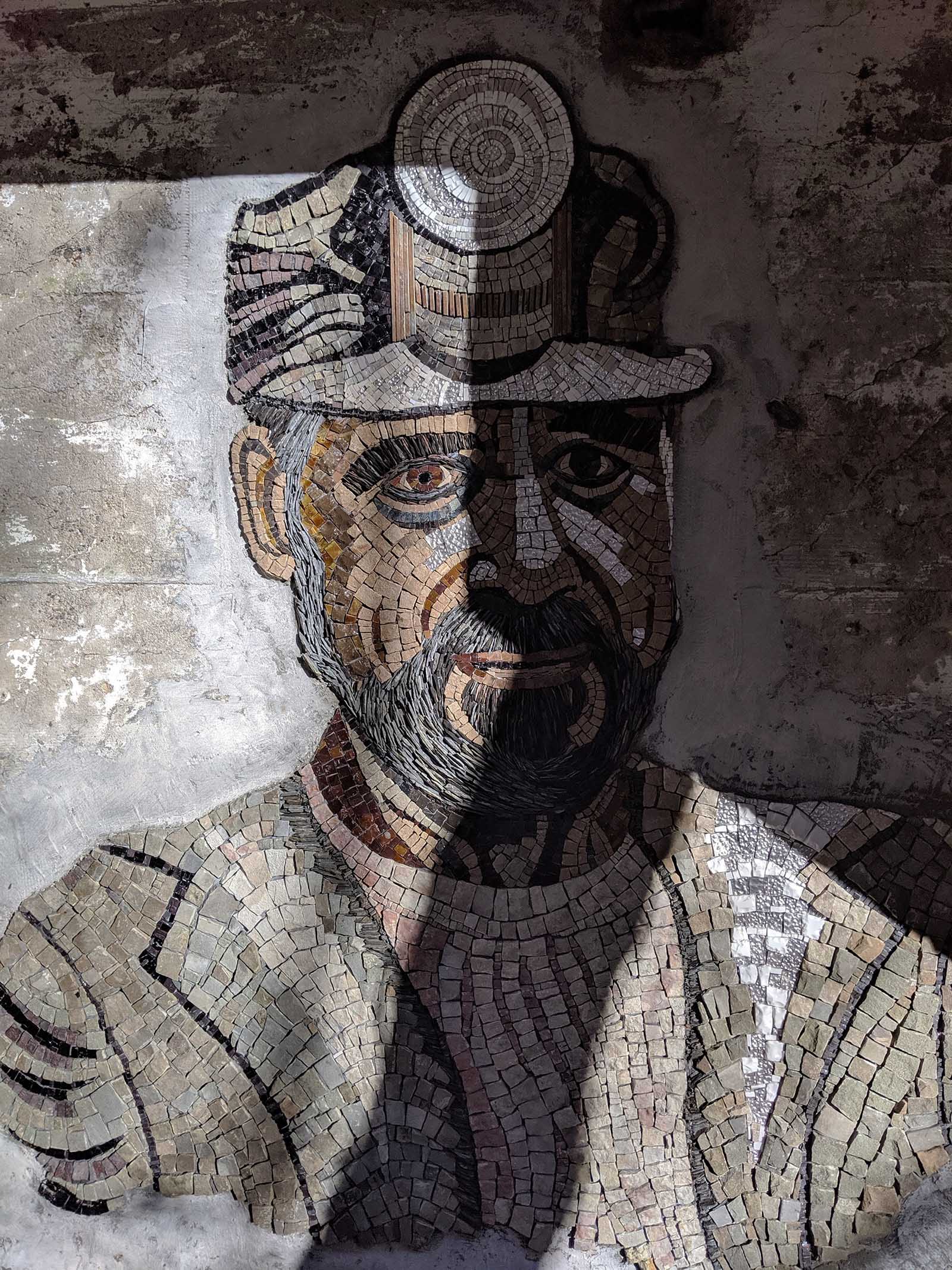 An earthtone mosaic of a miner with a lamp on this head with a beard and wearing a jacket over a sweater. 