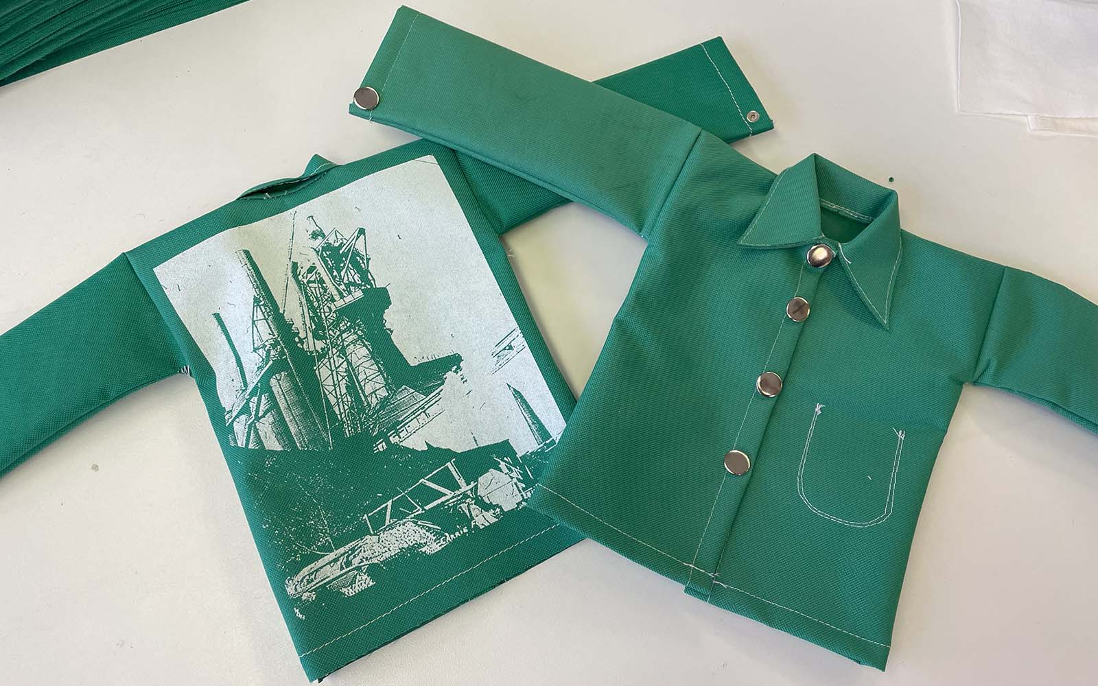 The back and front of a miniature workers jacket known as Greens. The back of the jacket has a silk-screened image of the Carrie Furnaces on it.