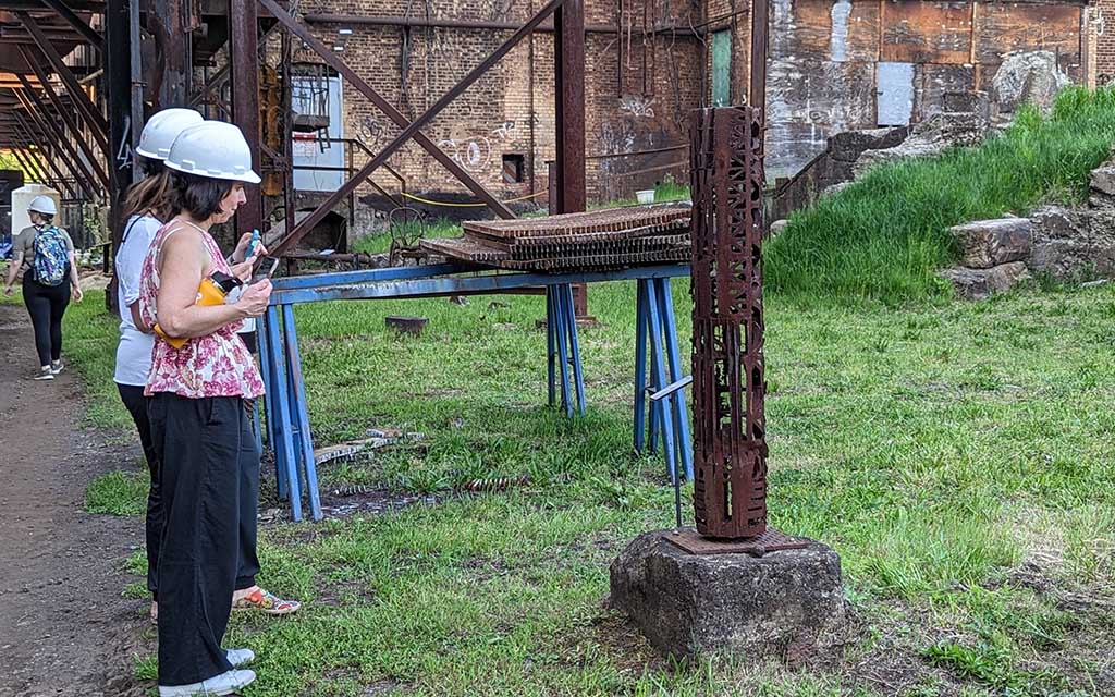 Two women in hard hats look at a rusty colored cylindrical sculpture.
