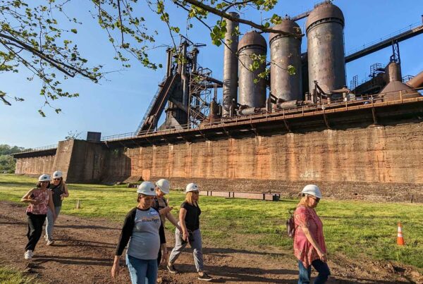 Educators in hard hats walk through the ore yard of the Carrie Blast Furnaces as the warm glow of a setting sun falls on the furnaces.