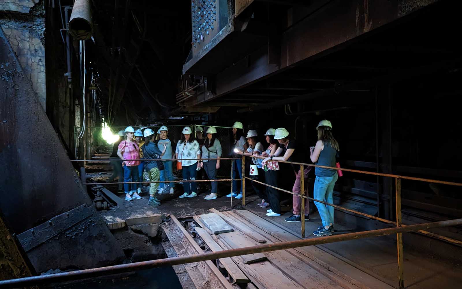 A group in hard hats tours the stock house, a dark space with rail tracks beneath the stocking trestle.