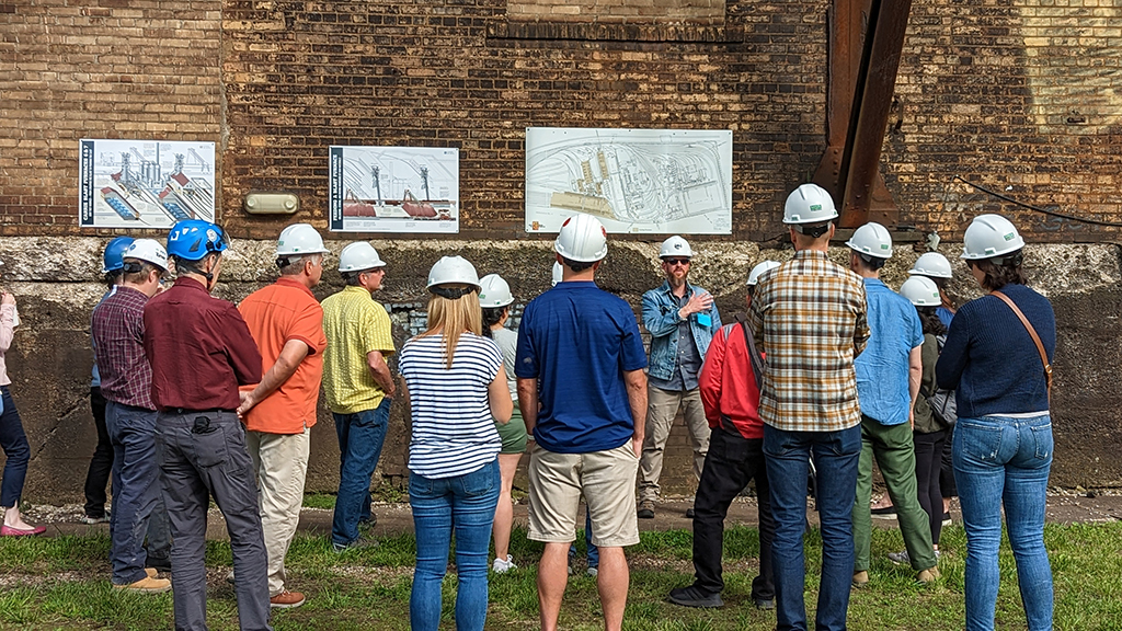 A group of mostly younger to middle age white folks in a hard hats gather around a tour guide who orients them to the space with a map behind him on a brick wall. 