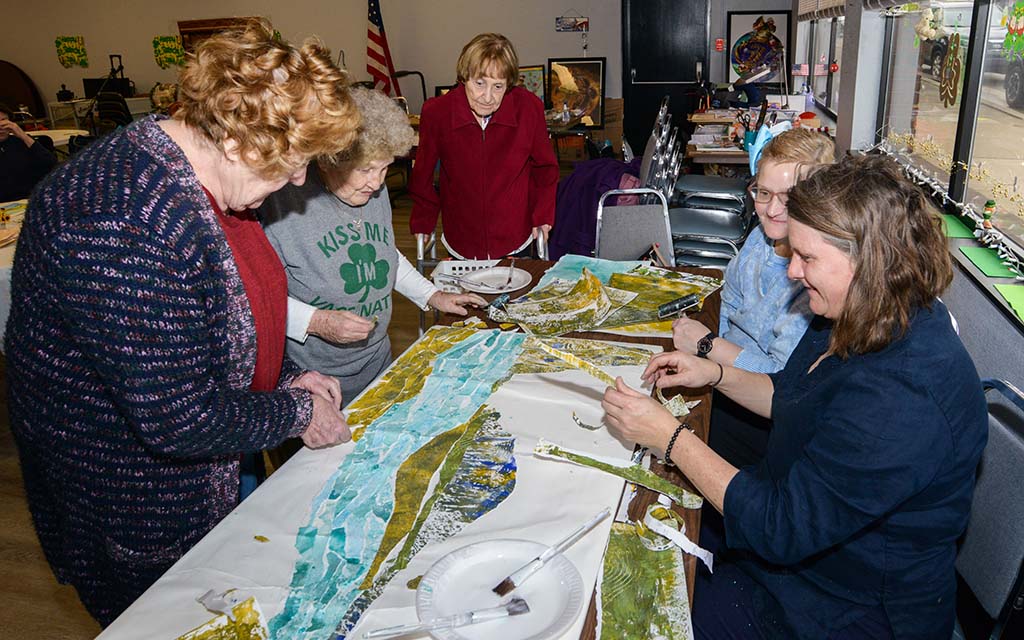 Five women gather around a table working on a collaborative art project.