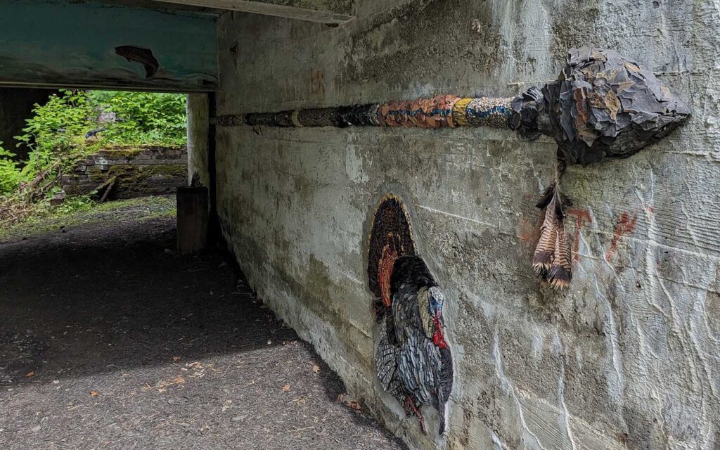 An artwork of an arrow stretches at least a dozen feet back along a concrete wall. It is applied to the surface, but the arrowhead itself rises up about eight inches from the wall. A flush image of a turkey is on the wall below it.
