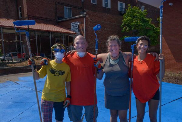 Three women and a man pose for a photo while holding paint rollers on extension poles. They are standing over a blue painted pavement that is clearly in progress.