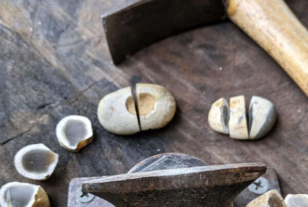 A still life of sorts showing a hammer a metal chisel anchored to a board, and several split rocks that have a gray center and white exterior.