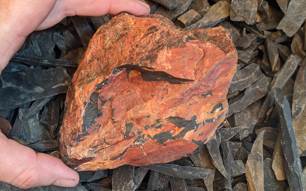 A reddish-orange rock that is nearly heart shaped being held between the middle finger and thumb on someone's hand, over a background of slivers of black stones.