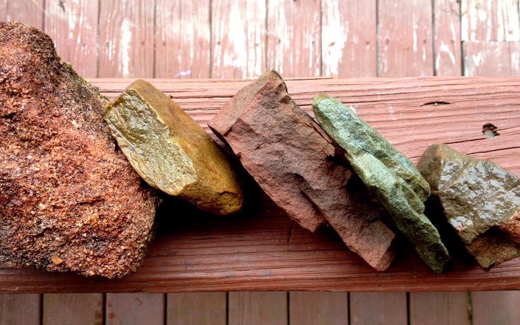Five rocks are arranged in a line to show their rainbow of colors—orange-red, yellow-tan, red-purple, green, and a mossy green.
