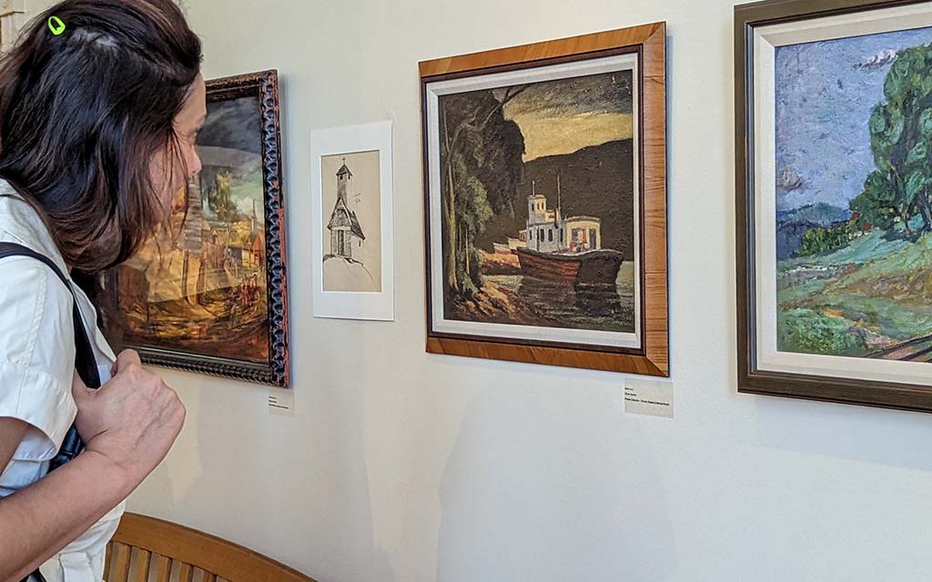 A woman leans in to view a realistic, pastoral painting. Another Van Gogh-esque impressionistic painting is adjacent to it.
