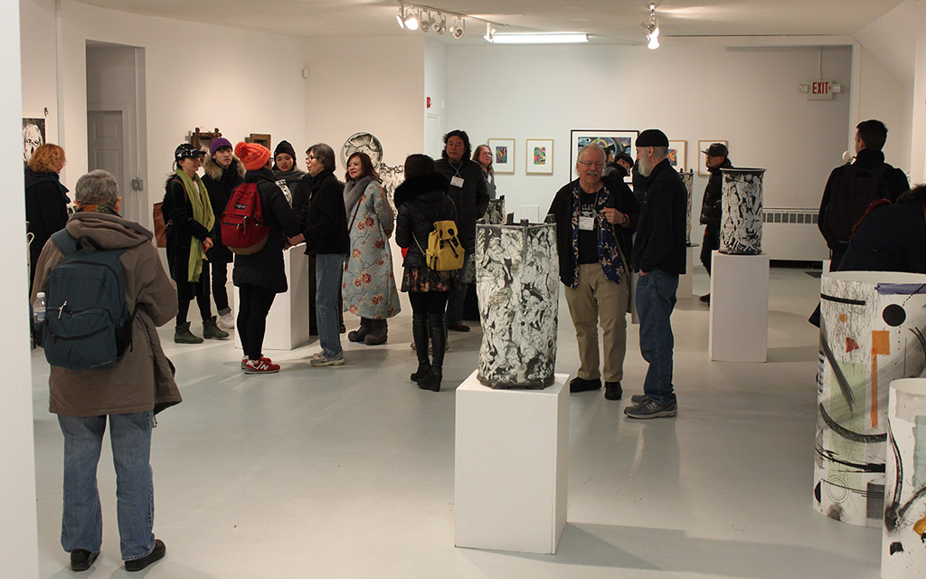 More than 15 patrons mil around in small groups in a white gallery space with large ceramic vessels on pedestals. 