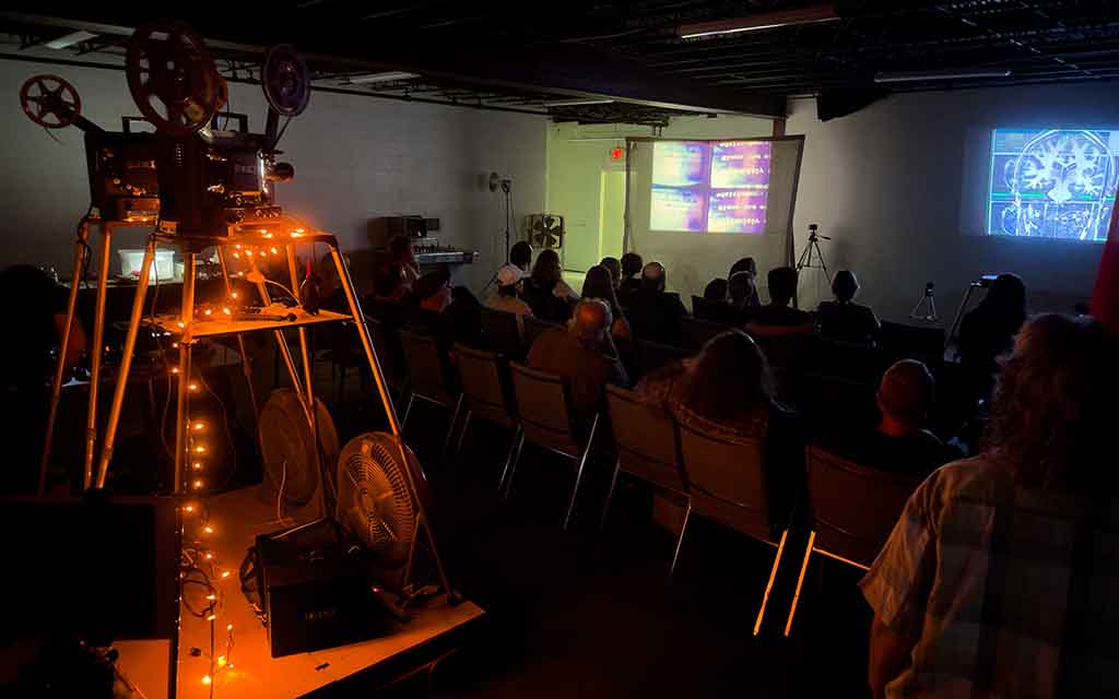 A film projector, draped in a strand of orange lights, projects an abstract image on a screen. In between the projector and the screen about thirty people sit in three rows of chairs viewing the film in near darkness.
