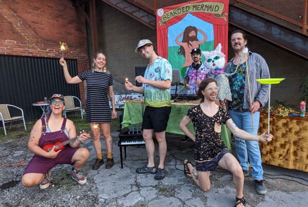Six eclectic, young white people pose for an image. Some hold sparklers, another has a puppet, one is spinning a plate, and there is a DJ in front of a circus-style bearded mermaid banner.