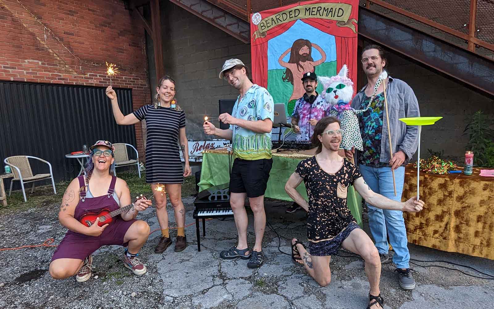 Six eclectic, young white people pose for an image. Some hold sparklers, another has a puppet, one is spinning a plate, and there is a DJ in front of a circus-style bearded mermaid banner.