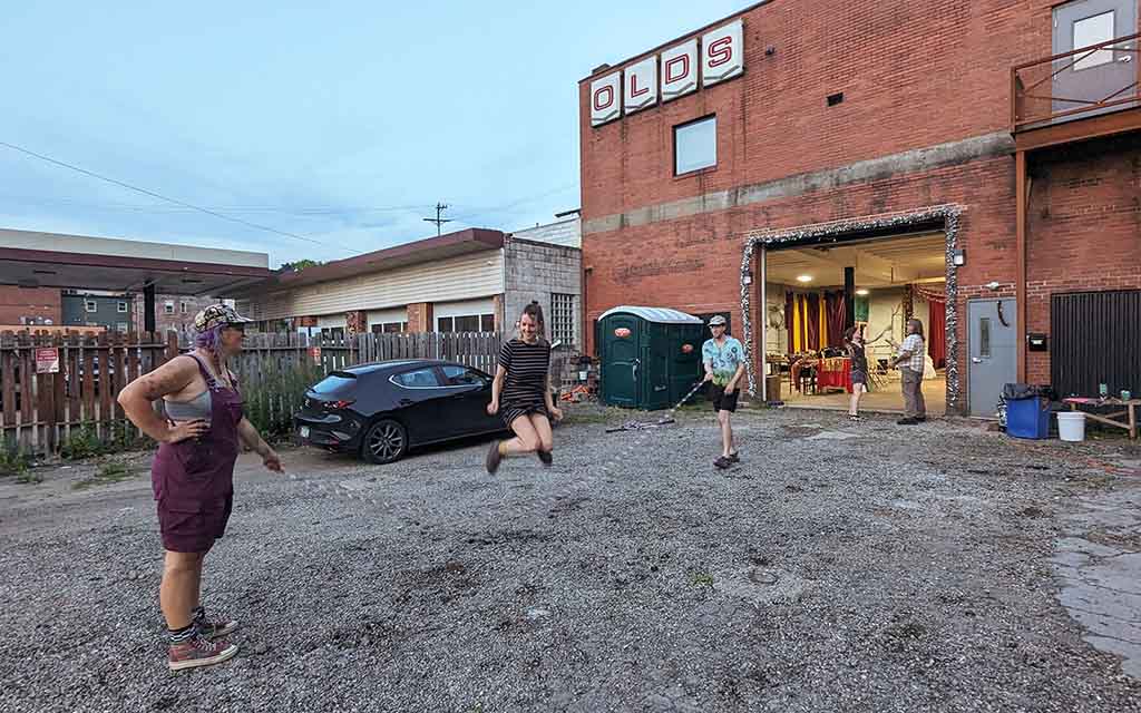 In front of a red brick building with a sign reading Olds, three people jump rope in the foreground what two other practice juggling in the background. 