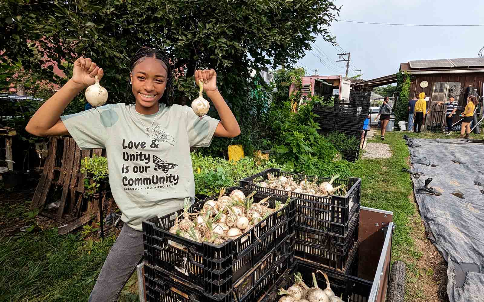 A black teen with small braids holds up two white onions she recently picked, standing in front of a cart full of baskets of onions. Her shirt reads "Love and Unity is our Community Braddock Youth Project."