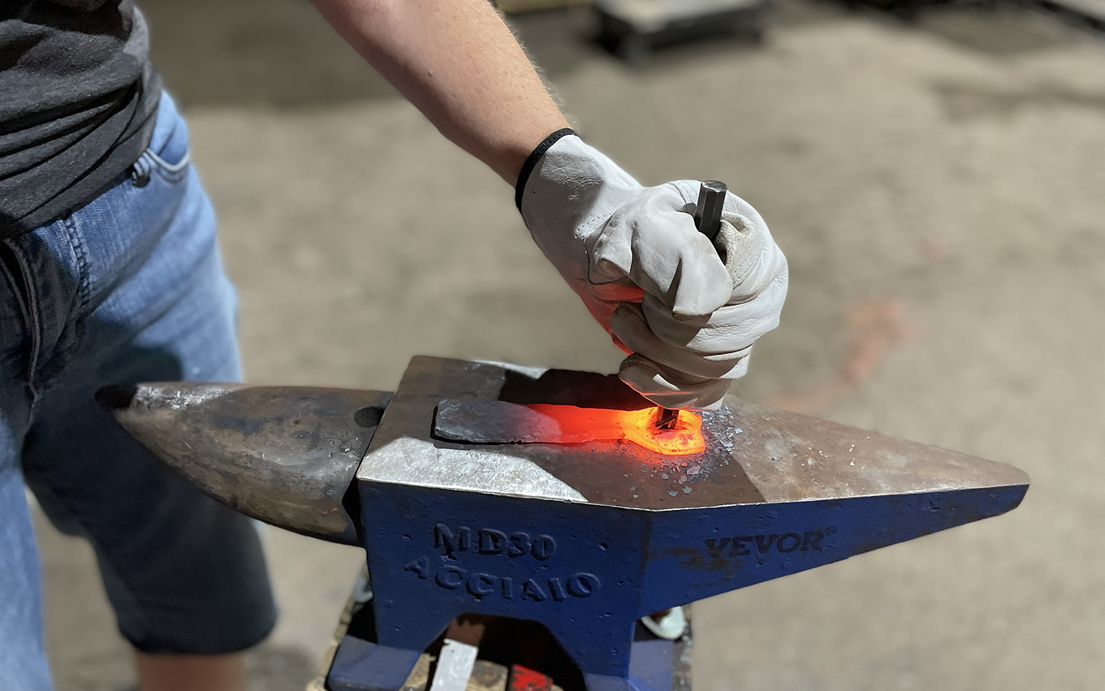 Close up image shows workshop participant hammering the chisel into red hot steel to open the bottle opener head.