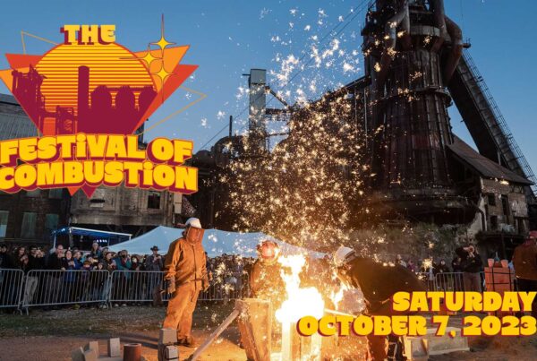 Sparks ascend towards the sky as a metal arts crew creates a performance iron pour in front of a crowd of people with the historic blast furnace behind them.