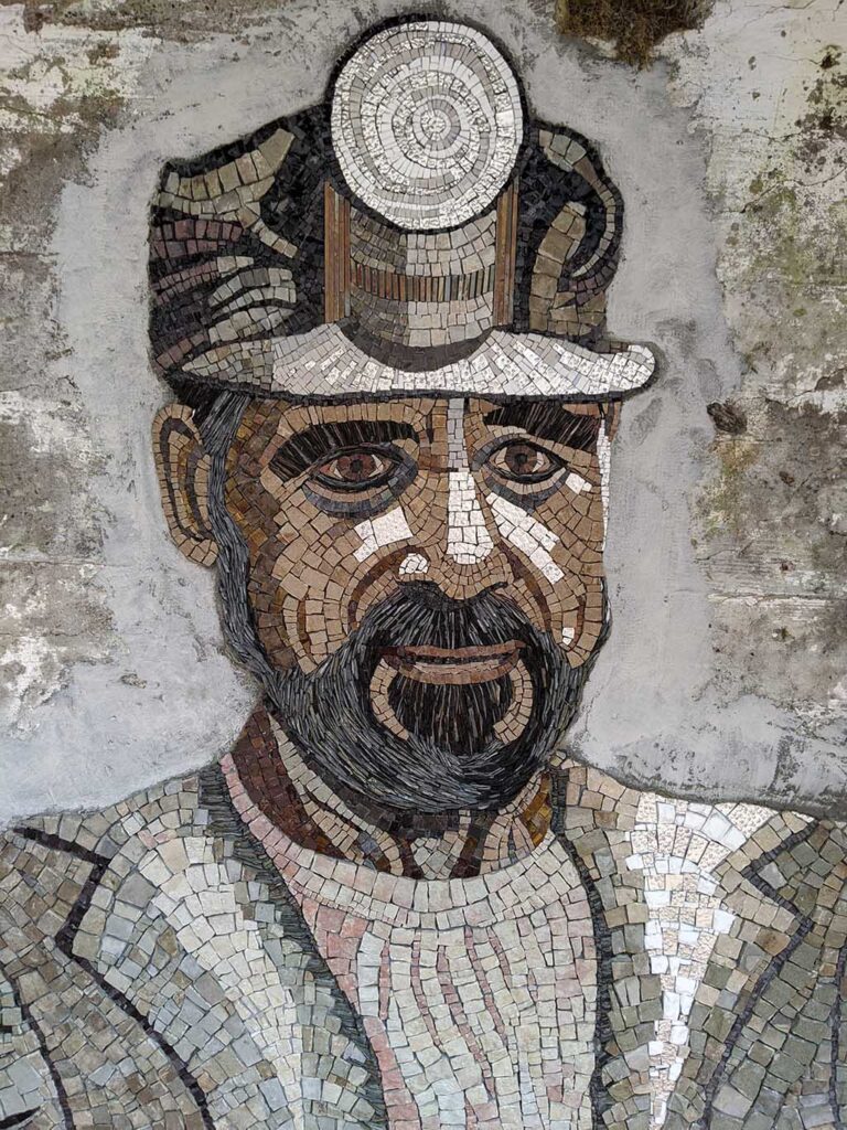 A mosaic image of a miner with a light on his forehead wearing a jacket with lapels. The lines of the image contour the shape of this face, goatee, and curvature of his chest.