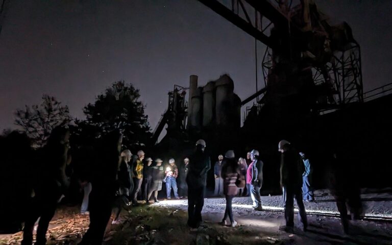 A group stand in the ore yard with flashlights with the silhouette of the furnace behind them.