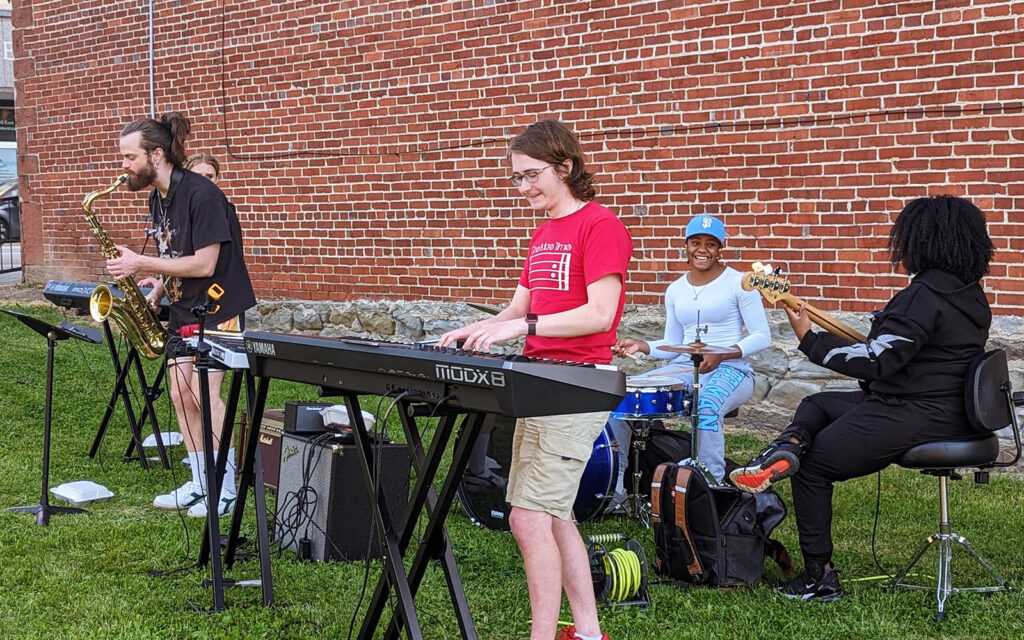 Teens perform in a grassy lot with a brick wall behind them. 