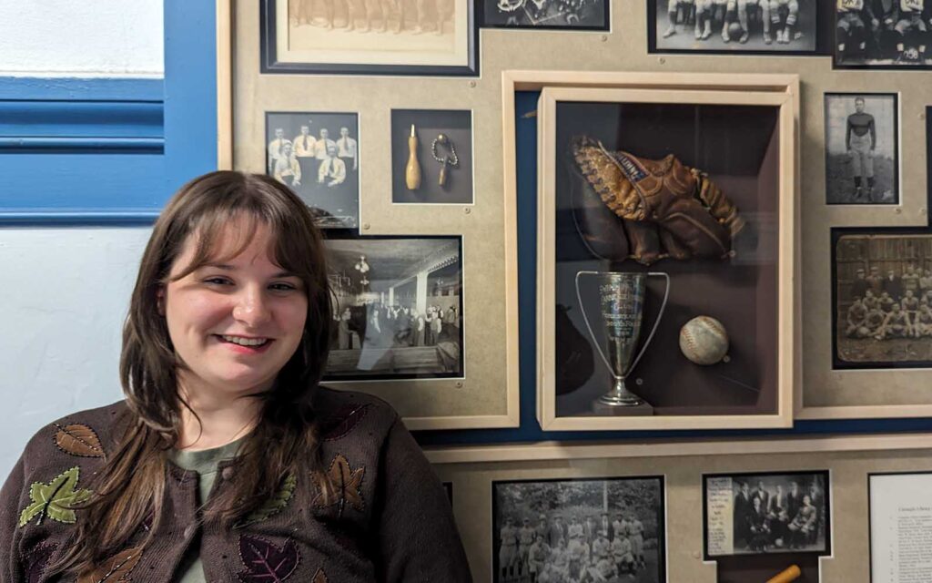 A younger white woman with bangs smiles in front of a wall of old photographs and a shadowbox with a trophy and sports equipment in it.