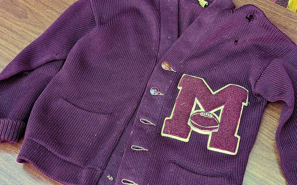 A purple sweater with holes near the left shoulder and pockets near the lower hem, boast the letter "M" with an image of a football sewn to the garment.