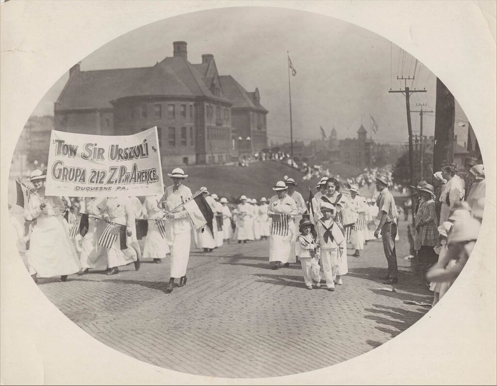 Dozens of people all dressed in white parade down the street in front of a large building. The ones in the front hold a flag, with text written in Polish.