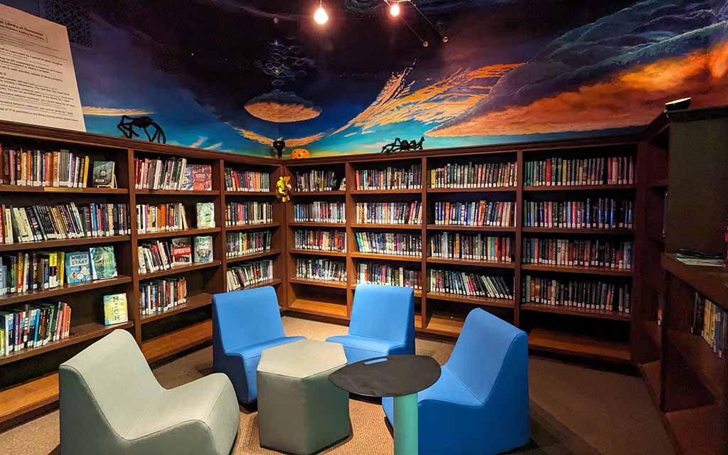 Four modern, low slung chairs are gather in the corner of a room with walls filled high with books. Above the shelfs is a mural of clouds and sky. 