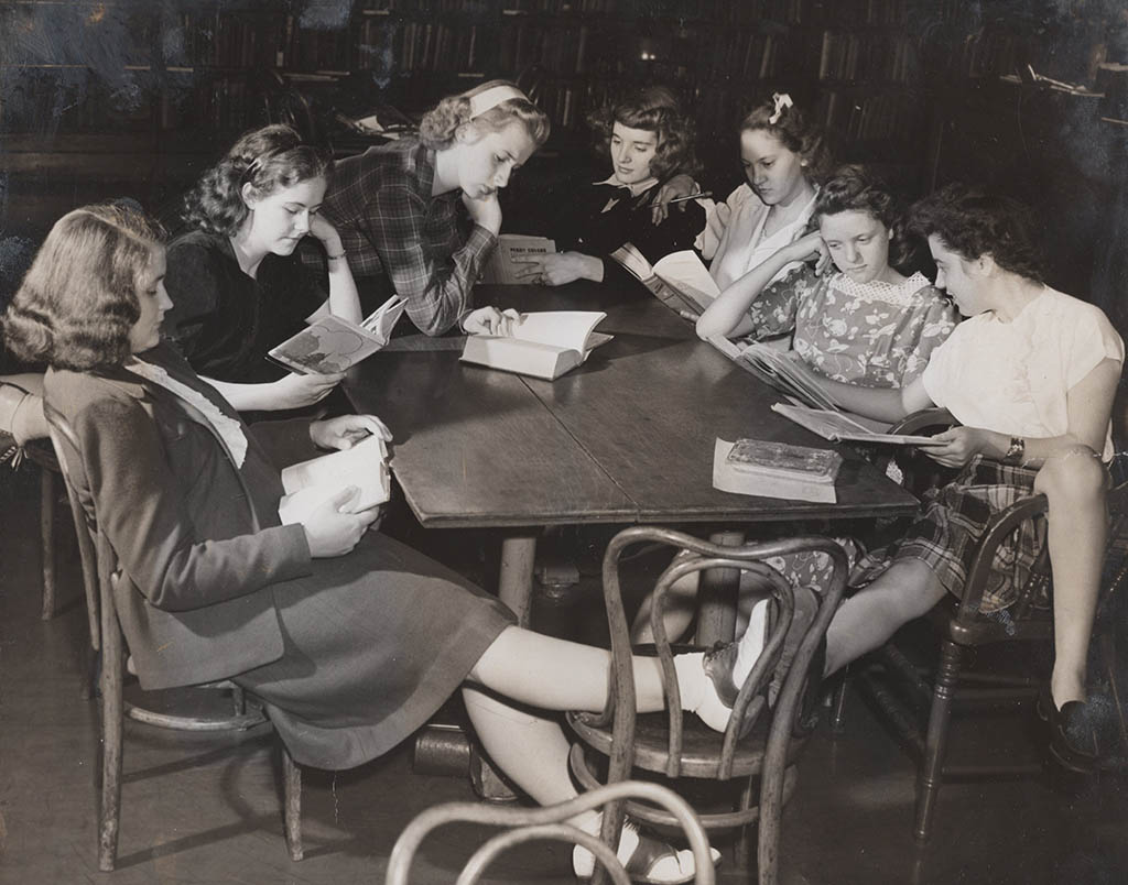 Six teenage girls in knee-length dresses and skirts gather with various reading material around a table in this black and white photograph.