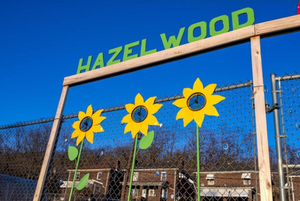 Art installed on a fence surrounding the Hazelwood Community Garden depicts three sunflowers below the word Hazelwood.