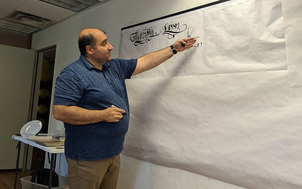 A somewhat rotund man of Turkish decent points a board with a drawing on it. It also reads "Calligraphy Lesson"
