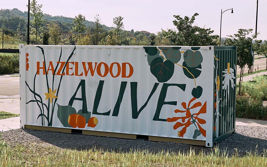 A shipping container is painted white with the word Hazelwood in orange, the word Alive in green, and foilage painted in orange, green and yellow.