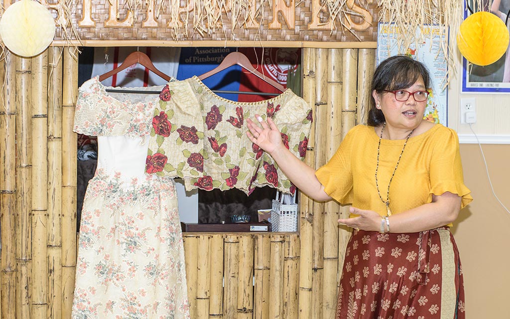 A Filipino woman stands and gestures towards two garments that are hanging behind her.
